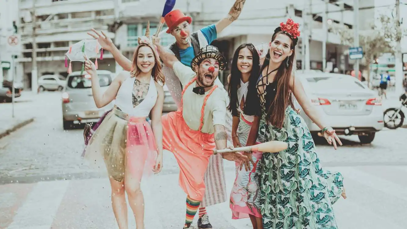 Dois homens vestidos de palhaço e três mulheres com roupas de Carnaval sorriem para foto na rua