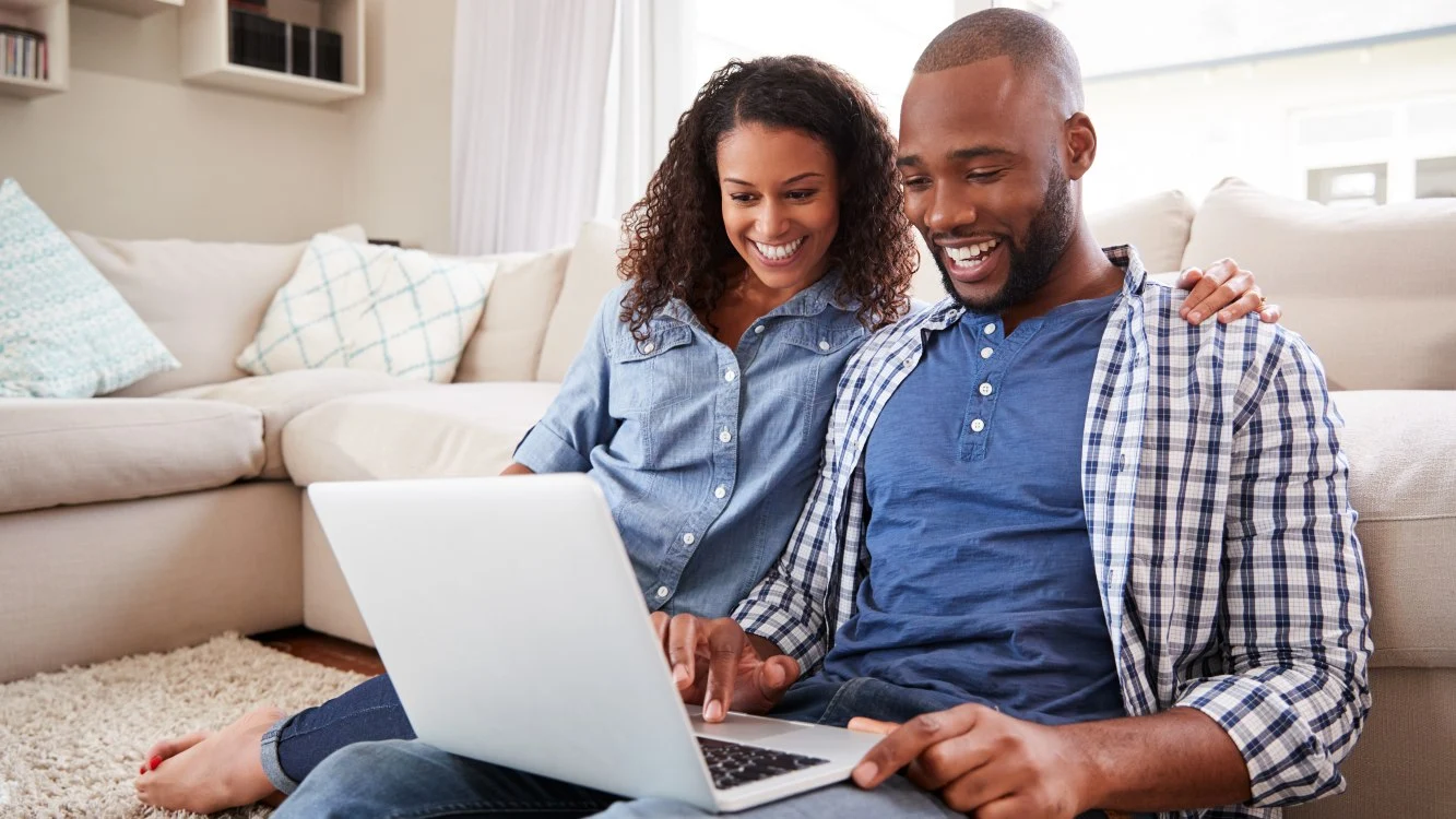 homem mexe no notebook e sorri. ao seu lado, uma mulher sentada sorrindo e olhando o notebook