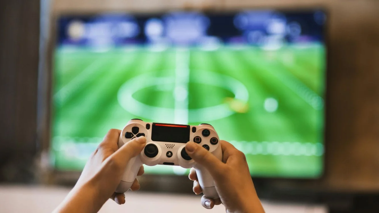 Duas mãos seguram um joystick. Ao fundo, monitor desfocado mostra partida de game de futebol