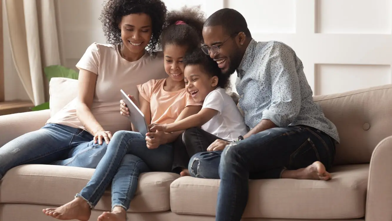 Dois adultos e duas meninas olham para a tela de um tablet na mão de uma das crianças. Os quatro estão sorrindo