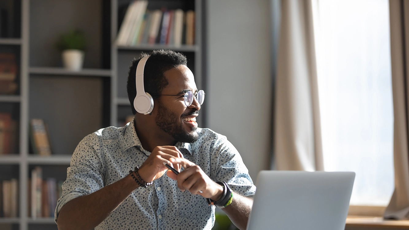 Homem sorrindo, usando fones de ouvidos sem fio, sentado de frente para um laptop.