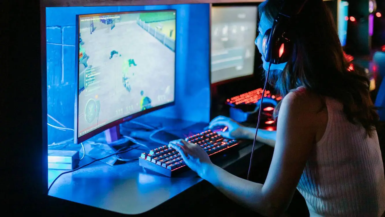 De perfil, garota com headset sorri em frente a um monitor. Na tela, há um jogo. O local está escuro e a única luz vem do monitor.