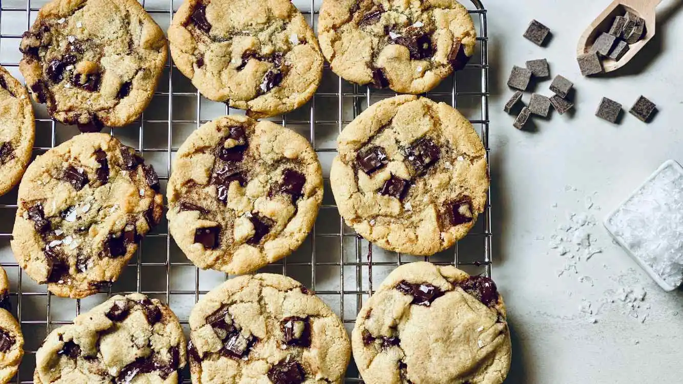 Fornada de cookies em cima de uma superfície. Ao lado, uma colher de pau com pedaços de chocolate espalhados por cima e ao redor dela