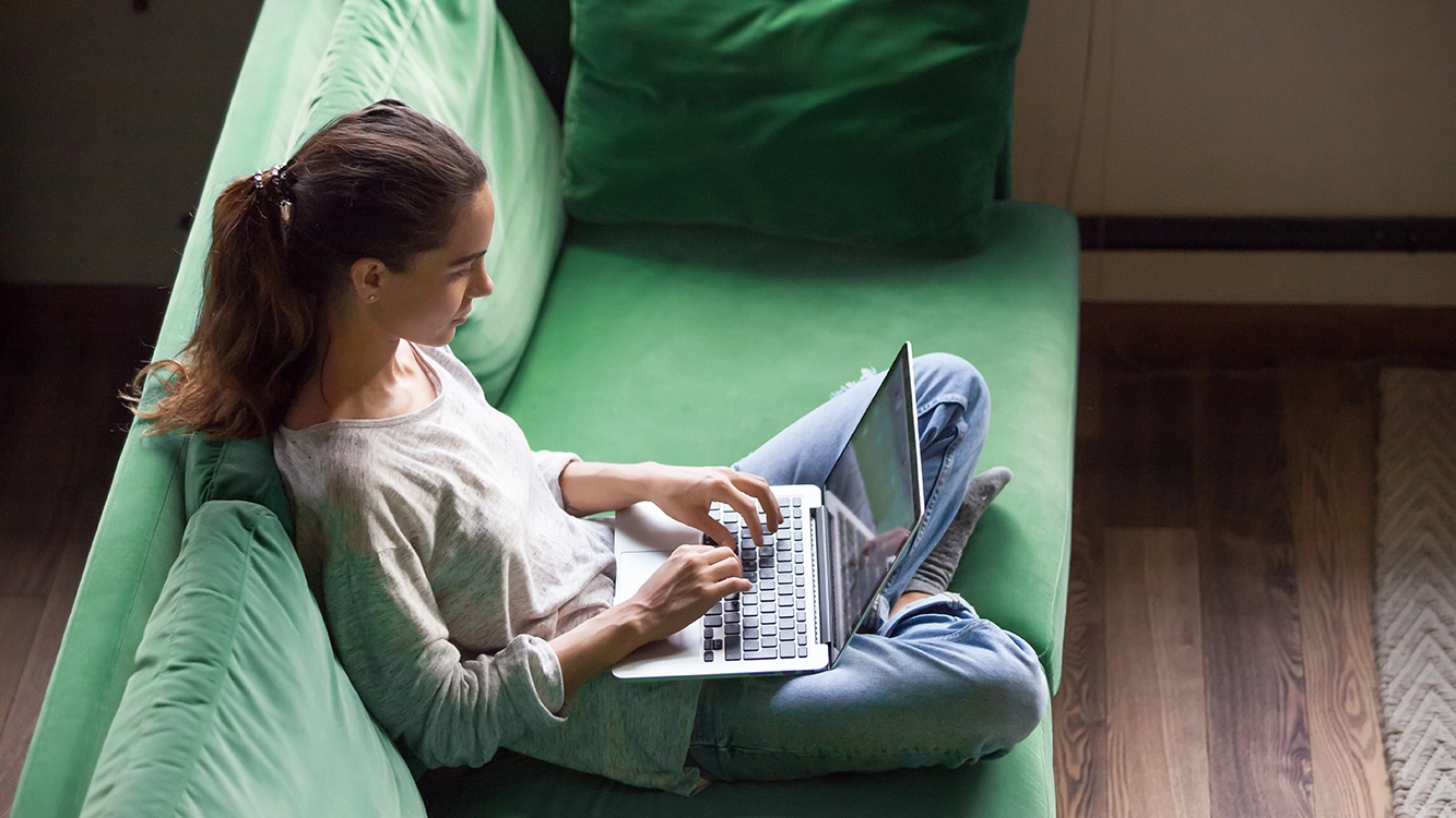Jovem mulher com um rabo de cavalo, sentada em um sofá verde, teclando no laptop.