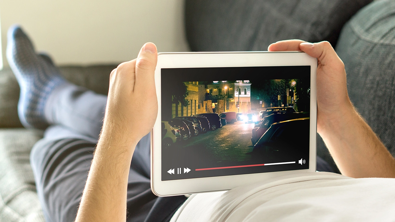 Homem deitado no sofá com um tablet nas mãos, assistindo a uma produção audiovisual.