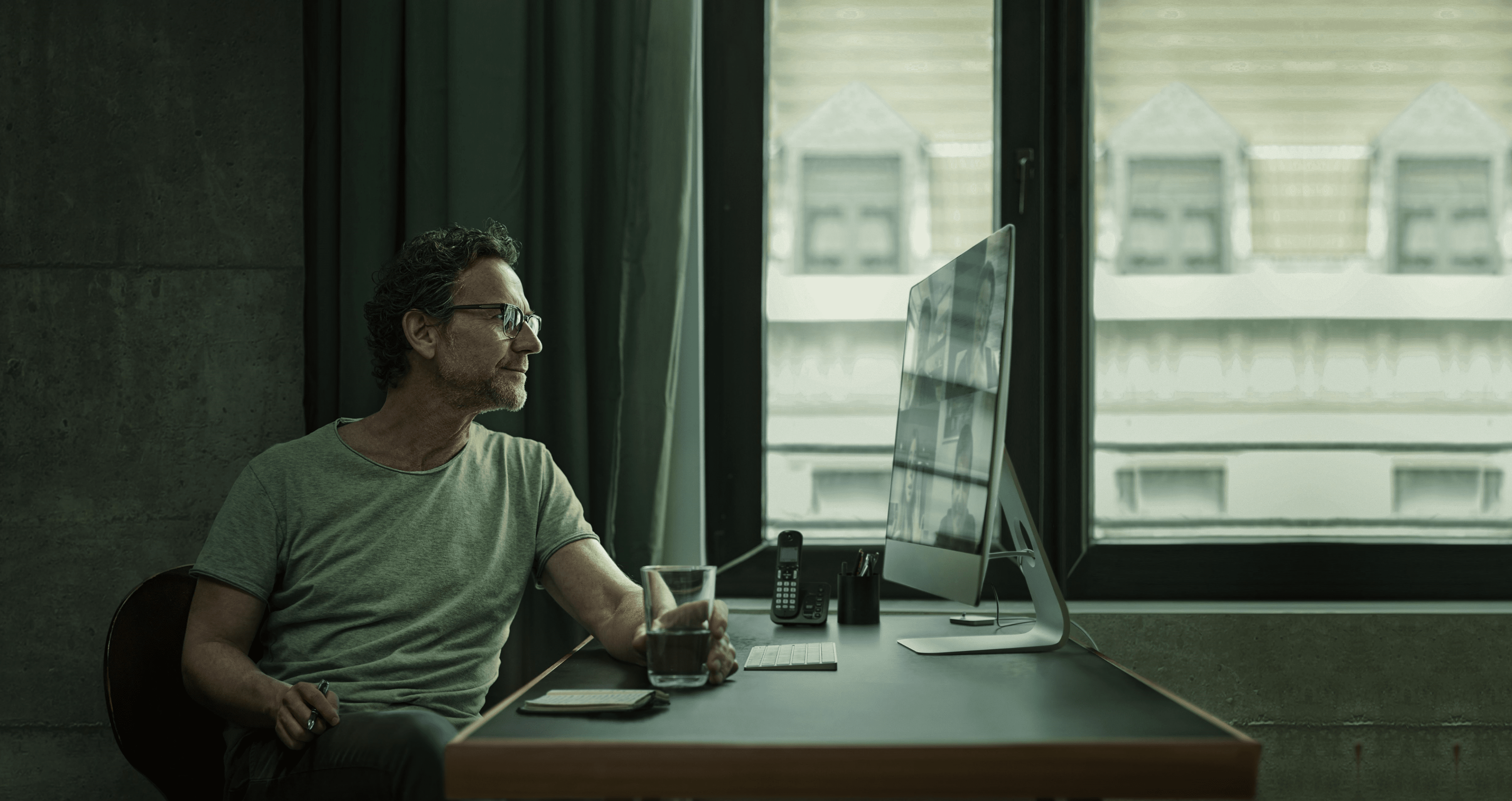 Homem branco em mesa de trabalho, segurando um café e olhando para monitor em videoconferência