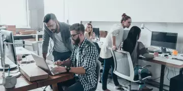 Um homem sentado e outro de pé estão trabalhando em um notebook com internet para empresas Oi Fibra. Ao fundo, outras três mulheres estão de costas também trabalhando.