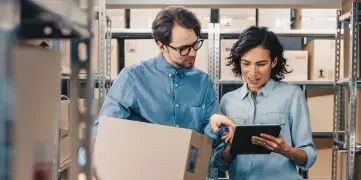 Na foto, um homem à esquerda e uma mulher ao seu lado conversam enquanto olham para a tela de um tablet. Eles estão trabalhando dentro do que parece ser o estoque de uma empresa.