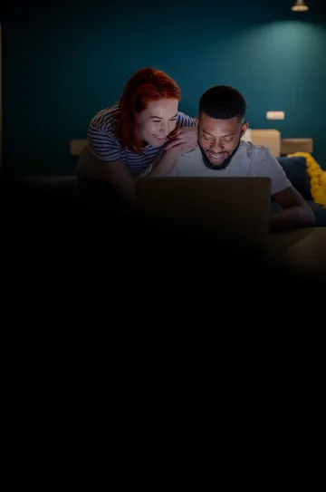 Mulher rindo, com as mãos nos ombros de um homem, observa a tela de um notebook que ele está usando