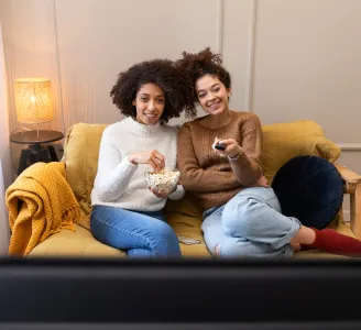 Duas mulheres, comendo pipoca no sofá da sala, assistem a série online