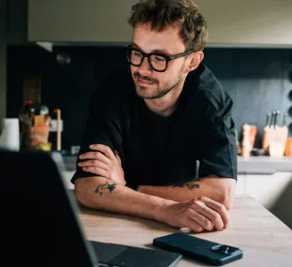 Homem de óculos, na cozinha de casa, usa computador e celular conectados à internet Oi Fibra