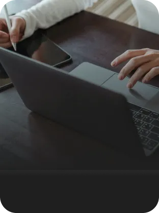 Pessoa escrevendo e utilizando um tablet sobre uma mesa de madeira, enquanto usa um notebook navegando com a banda larga Oi Fibra.