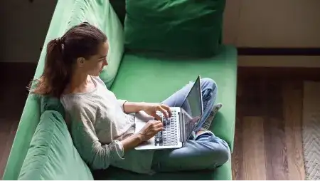 Mulher sentada no sofá teclando com o notebook no colo
