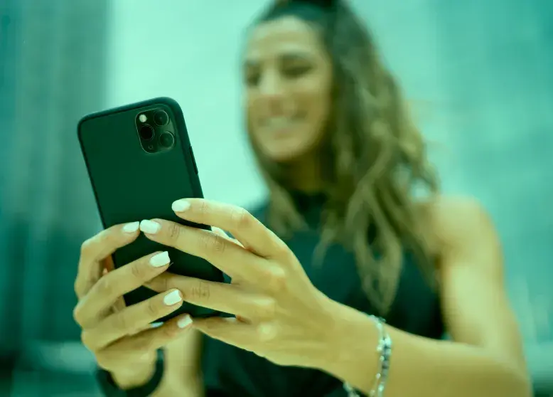 Foto mostra em primeiro plano a parte de trás de um celular nas mãos de mulher cujo rosto está em segundo plano e desfocado