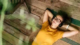 Mulher de camiseta laranja e óculos está deitada no chão com os braços sob a cabeça ouvindo música no fone de ouvido